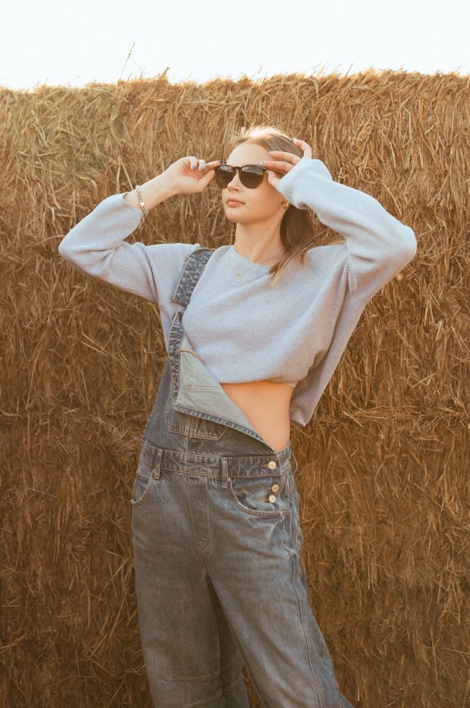 Person in denim overalls and cropped gray sweater poses against hay bale wall in sunglasses.