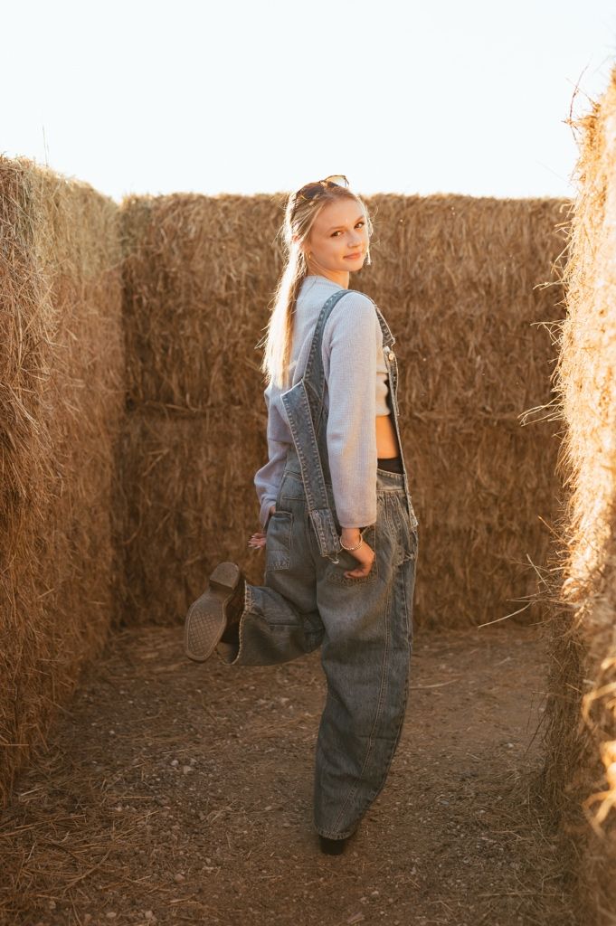 A person in denim overalls and gray sweater poses against a rustic hay bale wall in warm sunlight.