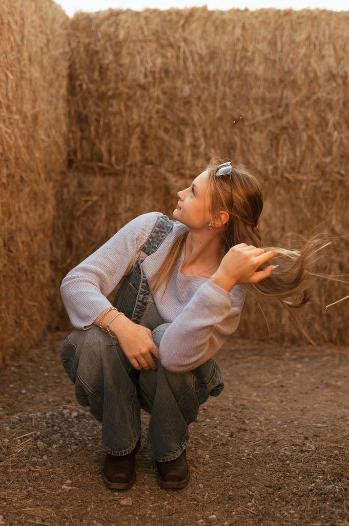 Artistic portrait of someone in denim overalls and white sweater sitting against a rustic hay bale wall at sunset.