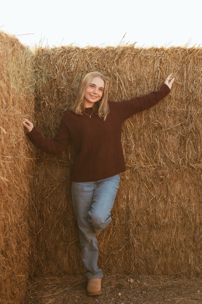Person in brown sweater and jeans poses playfully against rustic hay bale wall in outdoor setting.
