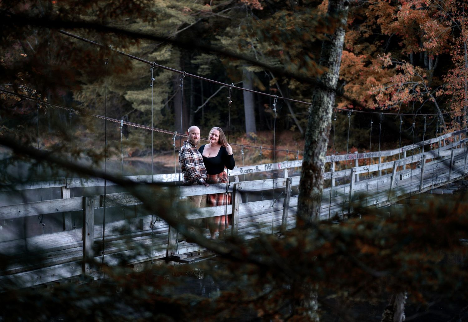 Two people walk along a wooden bridge surrounded by autumn foliage in a scenic outdoor setting.