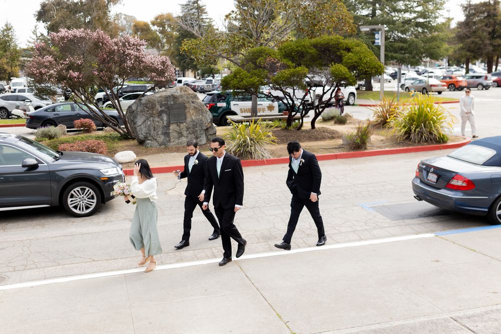 People walking through a parking lot with cars and trees visible in the background on a sunny day.