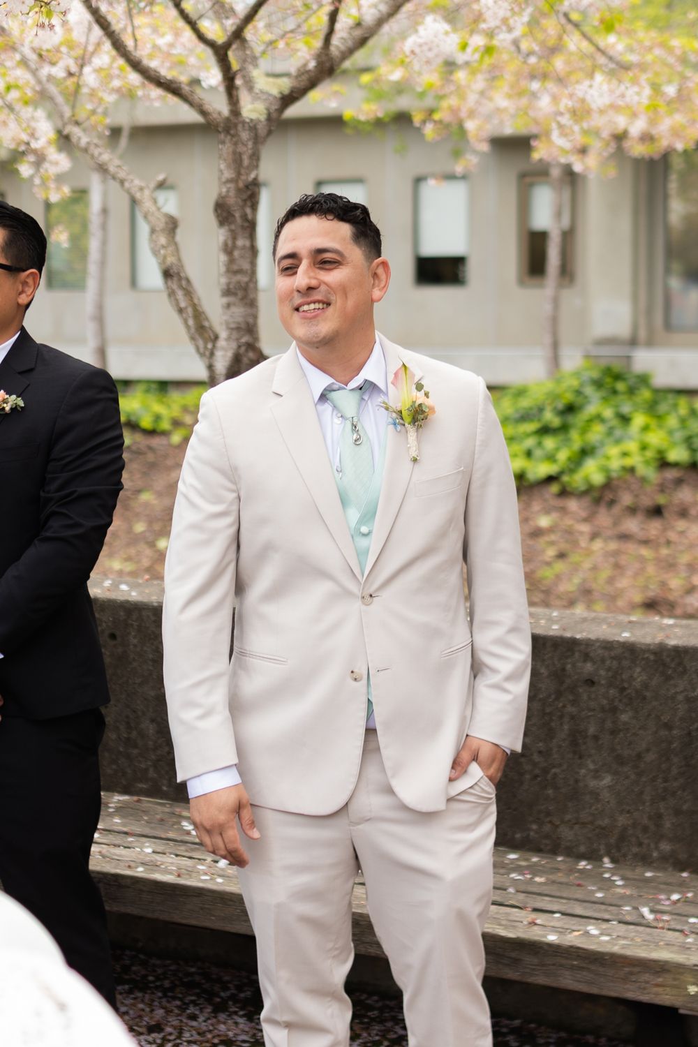 Wedding party member in a light khaki suit and mint green tie stands outdoors near trees and landscaping.