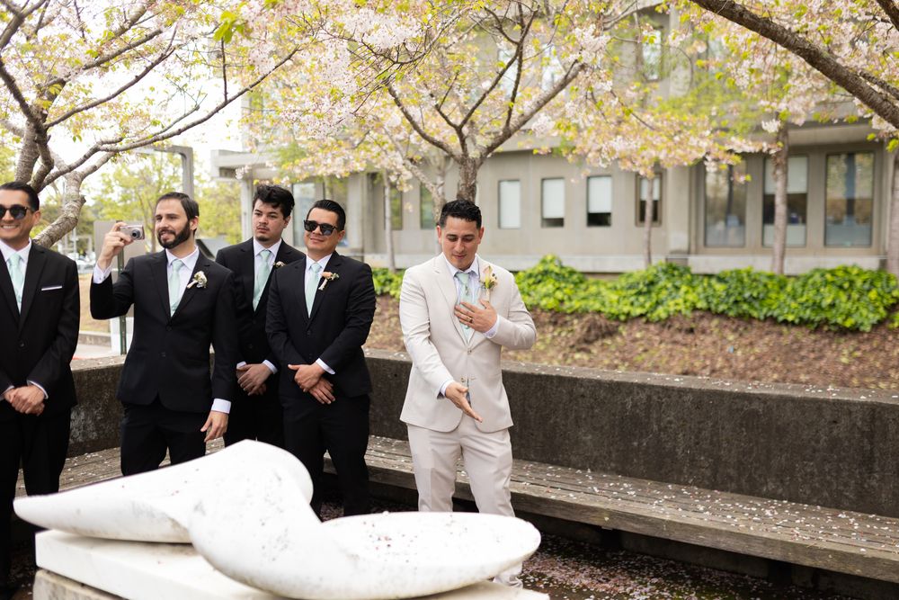 Wedding party members in black suits and one in white gather around a modern curved stone bench in an outdoor setting.