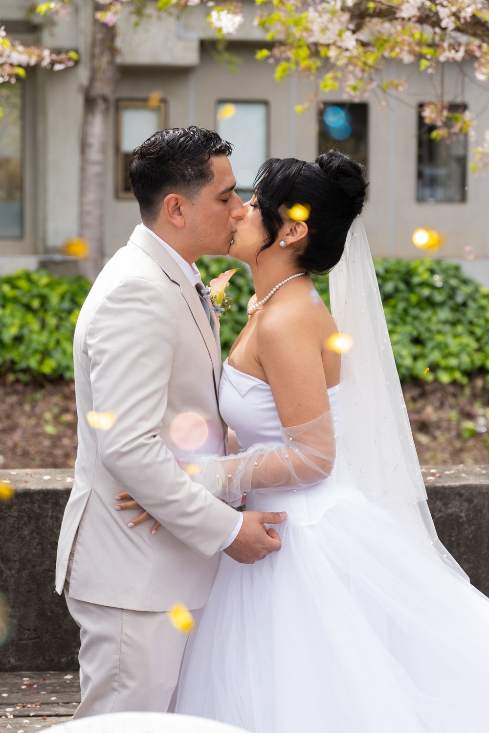 A romantic wedding couple in white attire share intimate moments in a garden setting with falling flower petals.