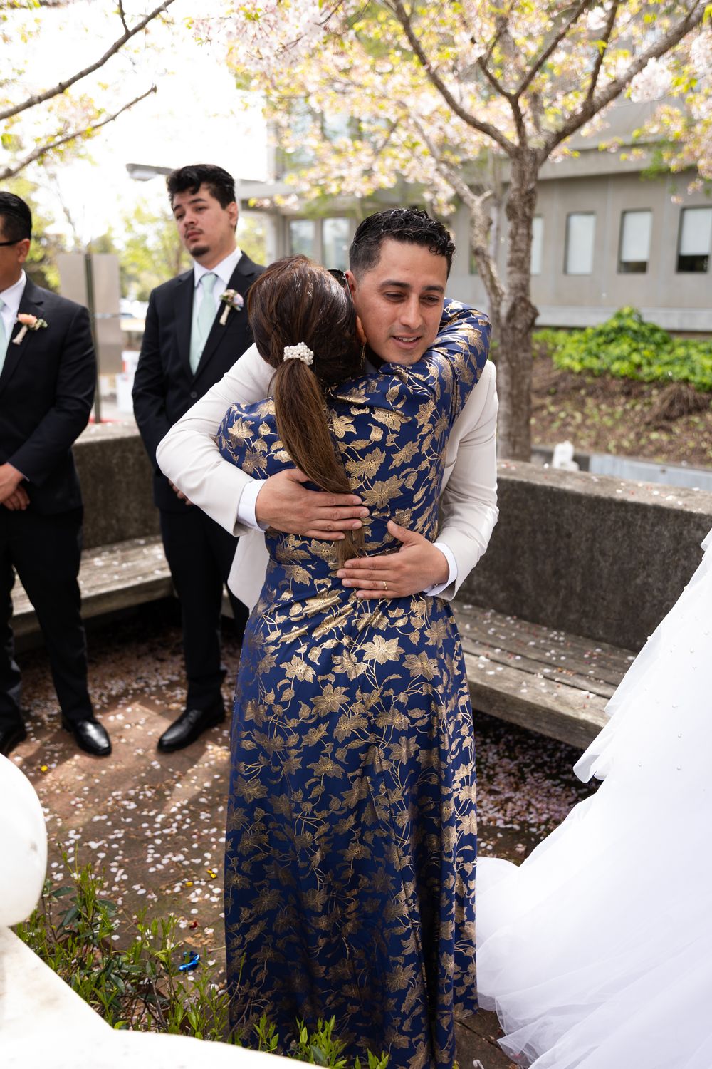 Wedding party members embrace in a garden setting with autumn trees and formal attire.