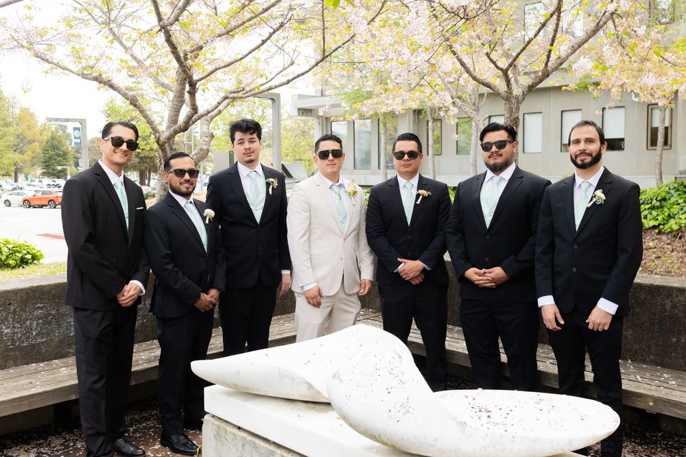 A wedding party in formal attire poses outdoors, with one member wearing light beige and others in black suits.