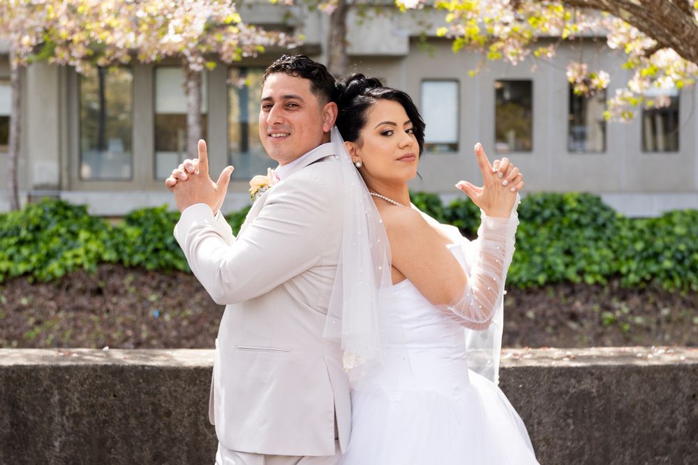A bride and groom pose back-to-back outdoors with playful expressions near a garden and building.