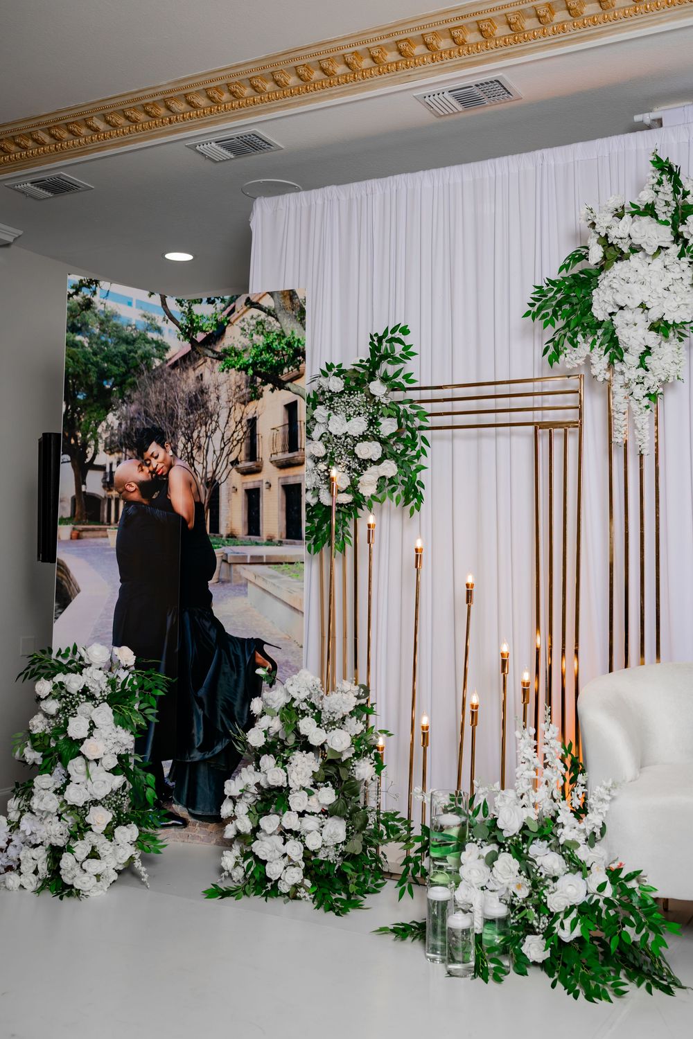 Elegant wedding ceremony backdrop with white drapes, gold geometric frame, white floral arrangements and greenery decorations.