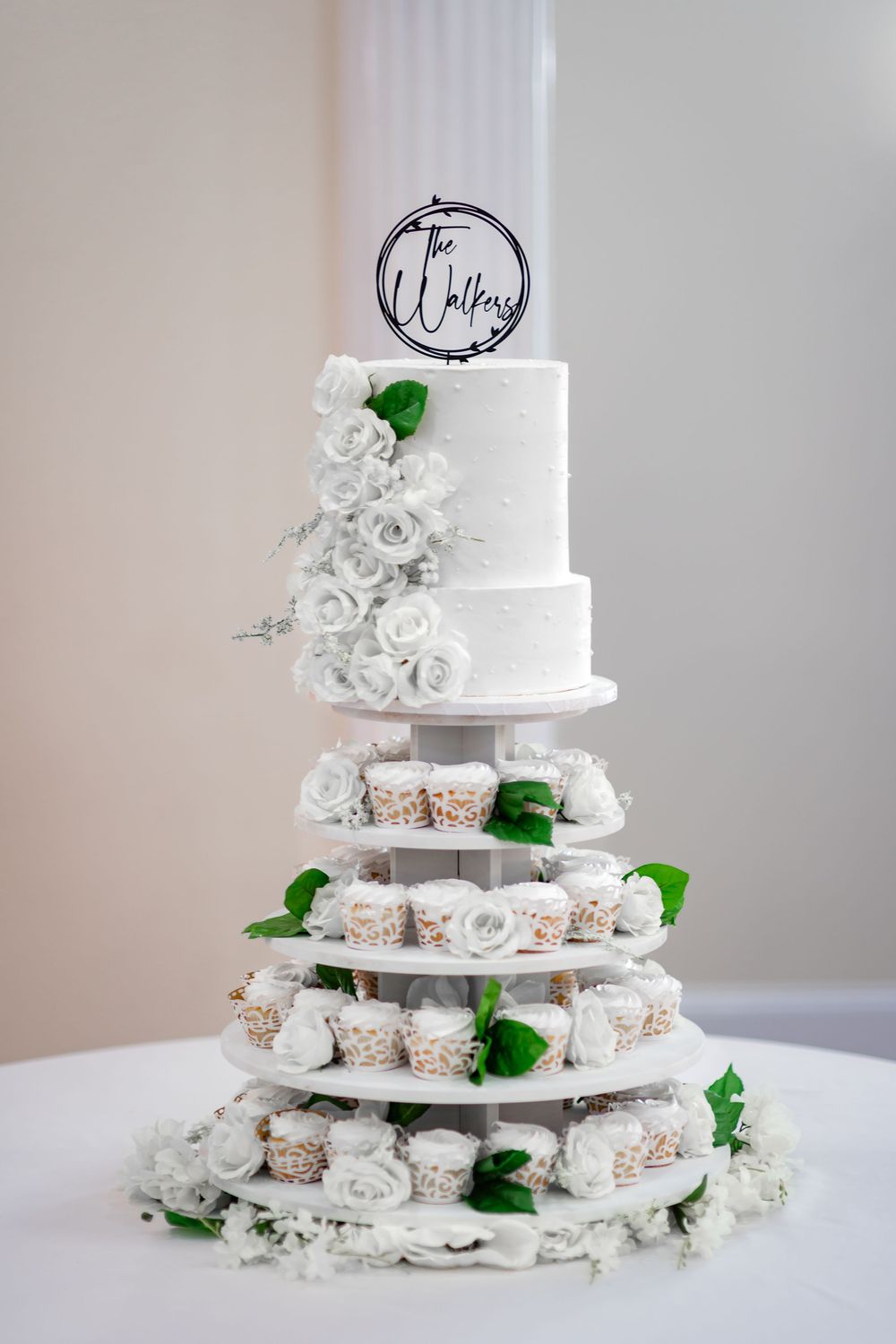 A tiered white wedding cake with roses and cupcakes displayed on an elegant silver stand against a neutral background.