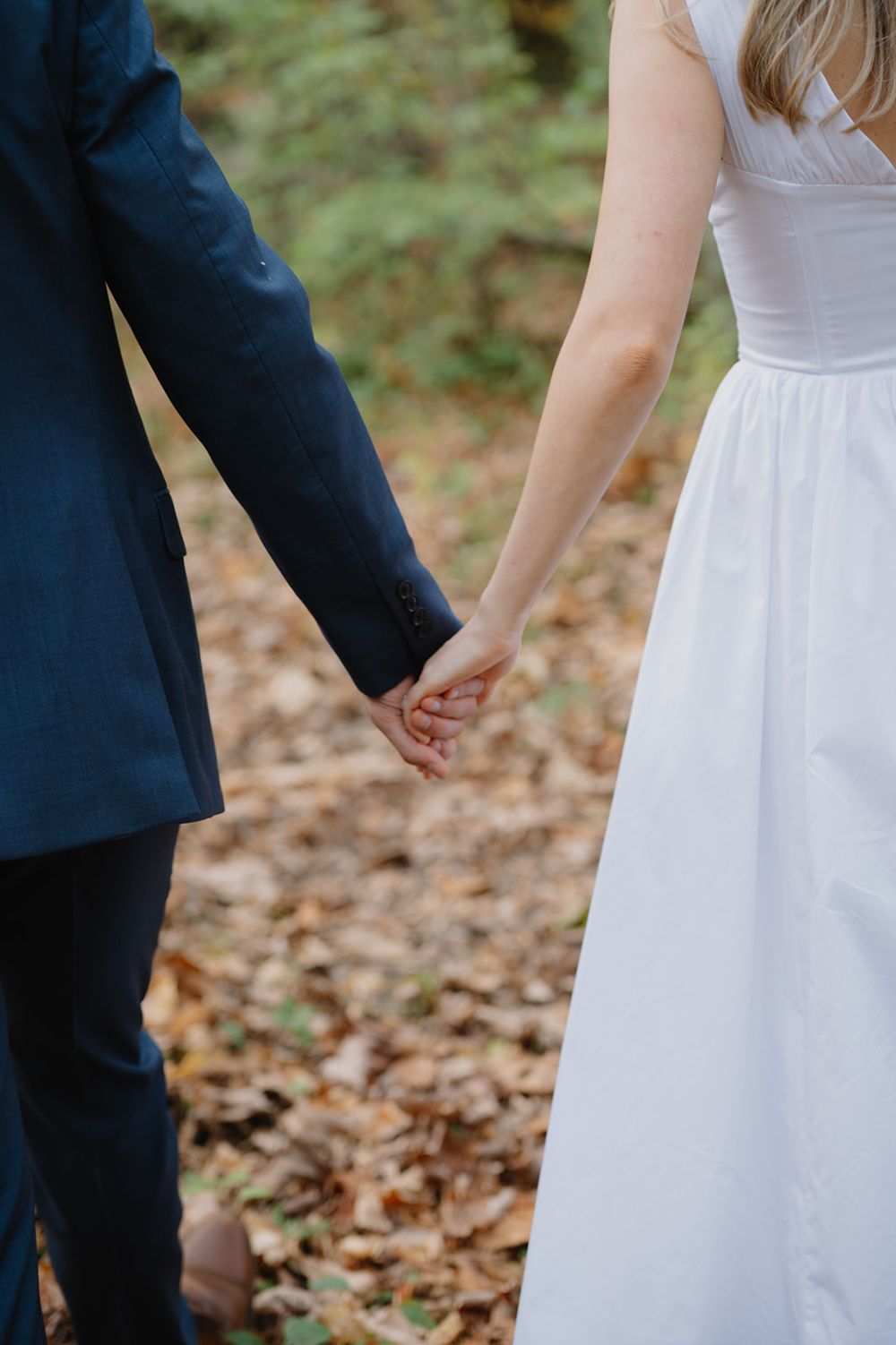 A couple in wedding attire holds hands while standing among fallen autumn leaves.