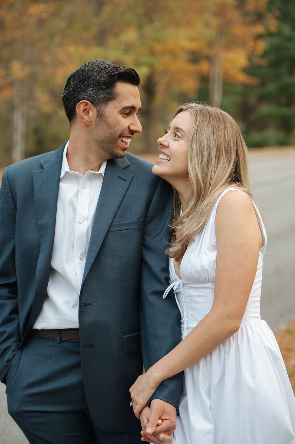 A couple in formal attire share a romantic moment outdoors with autumn foliage in the background.