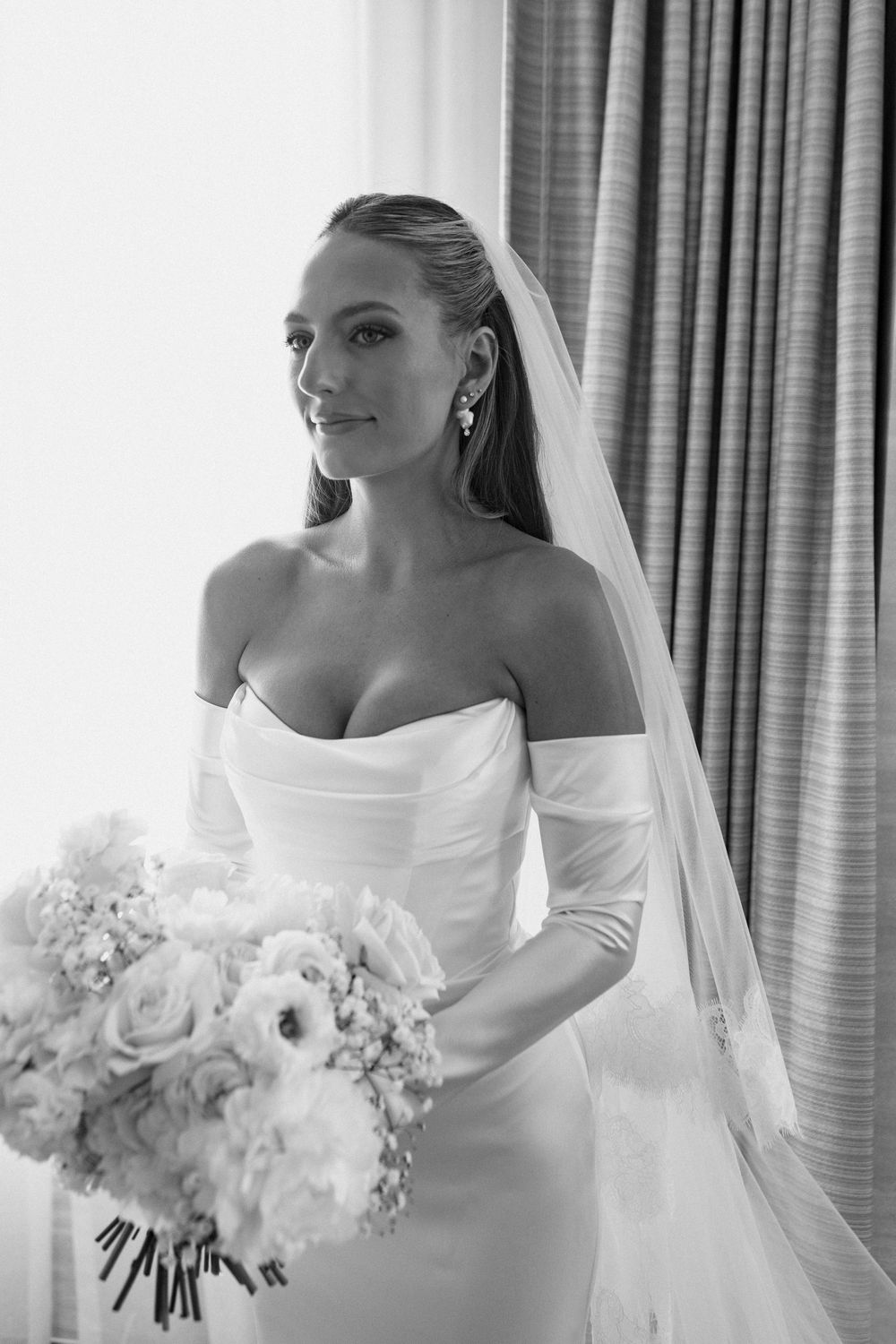A bride in an elegant white dress holds a bouquet of white roses and peonies against a curtained backdrop.