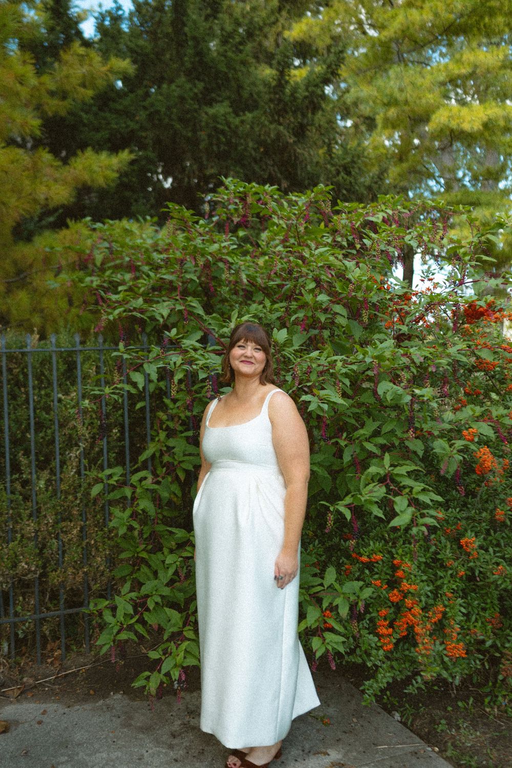 A person in a flowing white maxi dress stands in a garden setting with blooming red flowers and greenery.