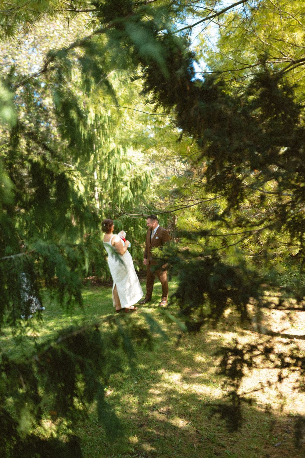 A figure in white clothing walks through a sunlit wooded area with dappled light filtering through green foliage.