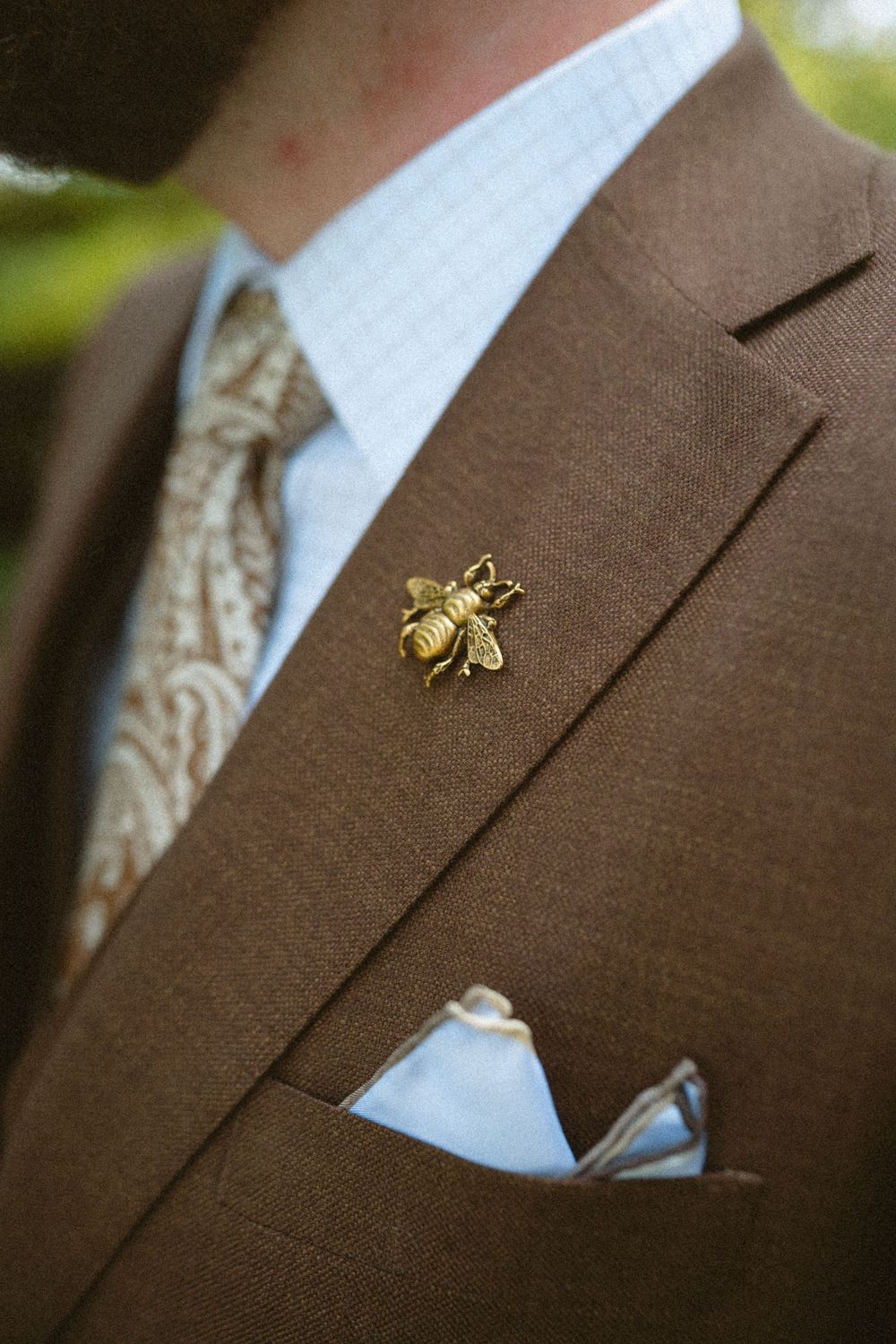 Gold bee lapel pin on brown suit jacket with white pocket square and patterned tie detail.