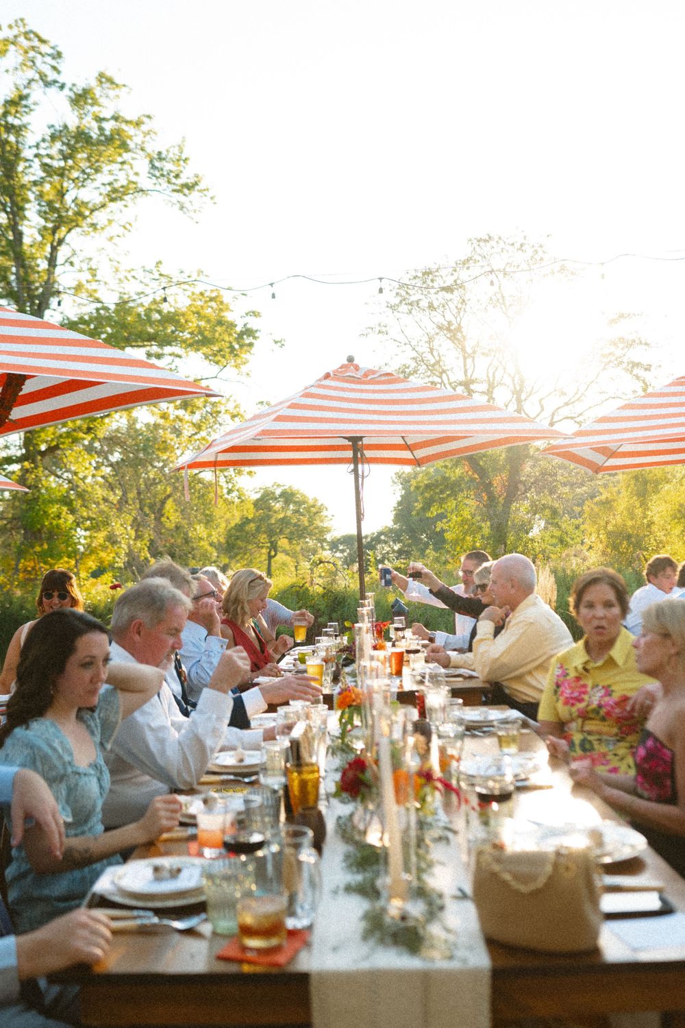Outdoor dinner party guests seated at long tables under red and white striped umbrellas during golden hour.