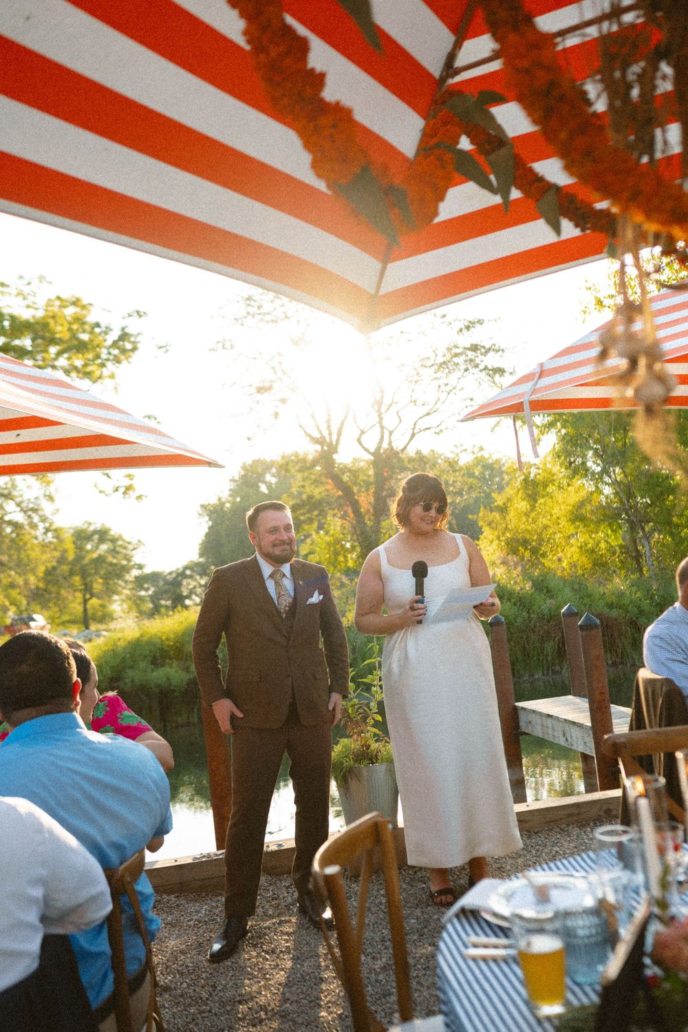 A wedding ceremony takes place under a red and white striped canopy during golden hour in an outdoor garden setting.