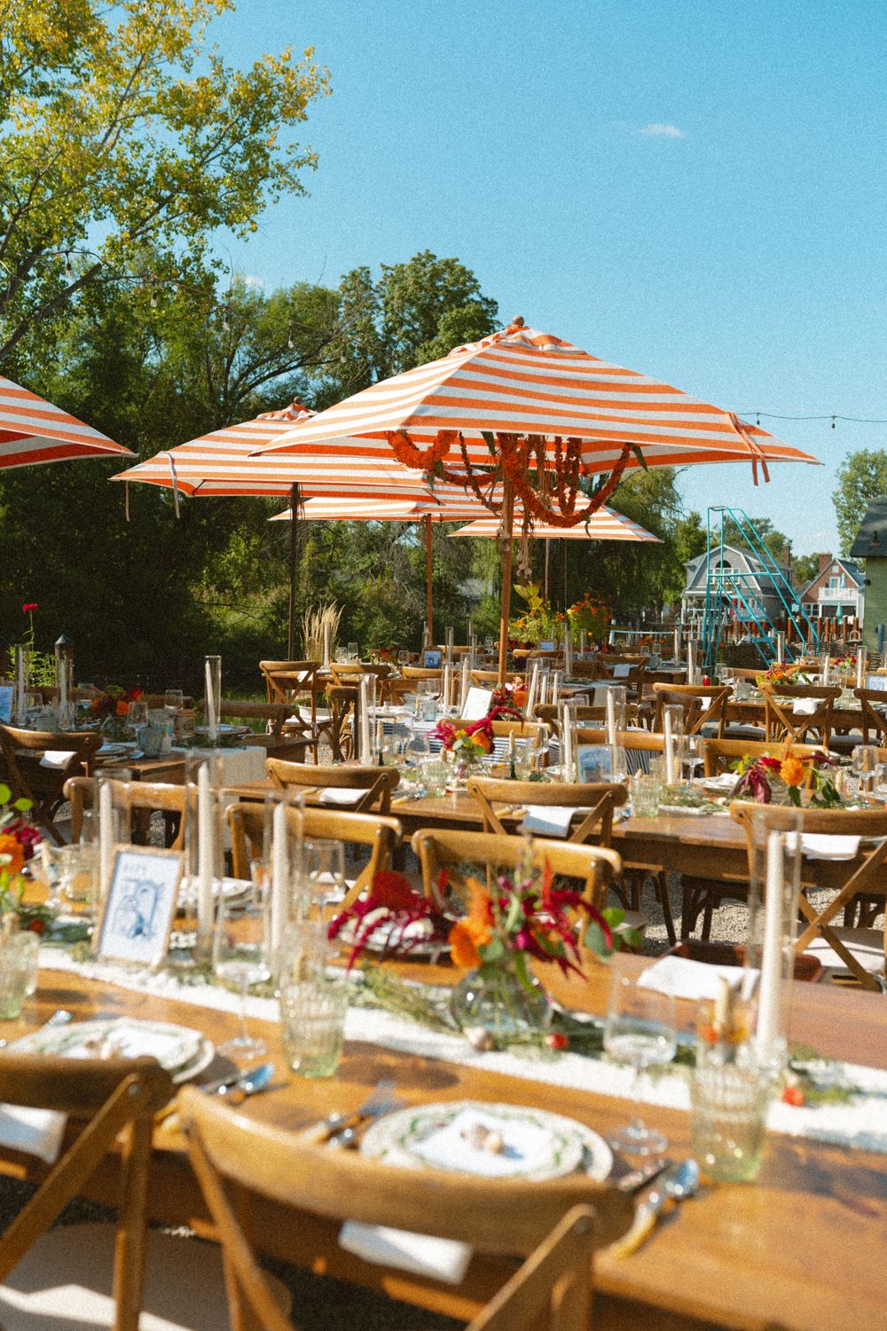 Outdoor restaurant seating area with wooden tables and orange striped umbrellas under blue summer sky.