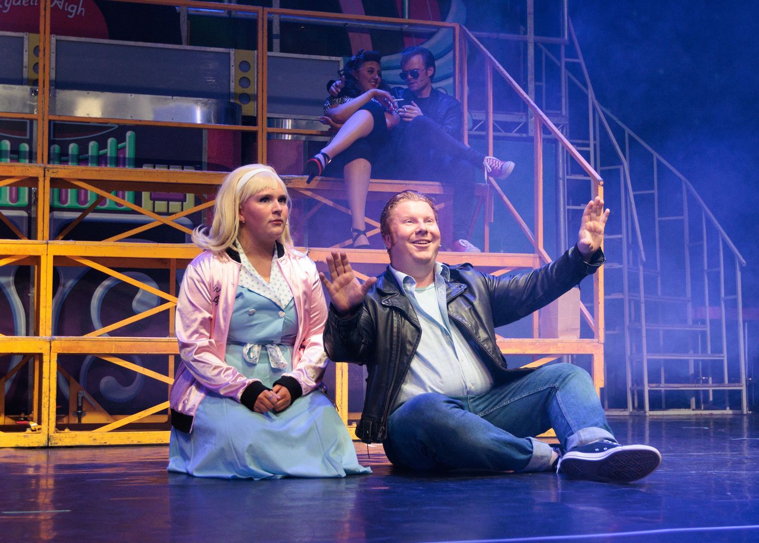 Performers act in a theatrical production with colorful stage lighting and scaffolding set pieces.