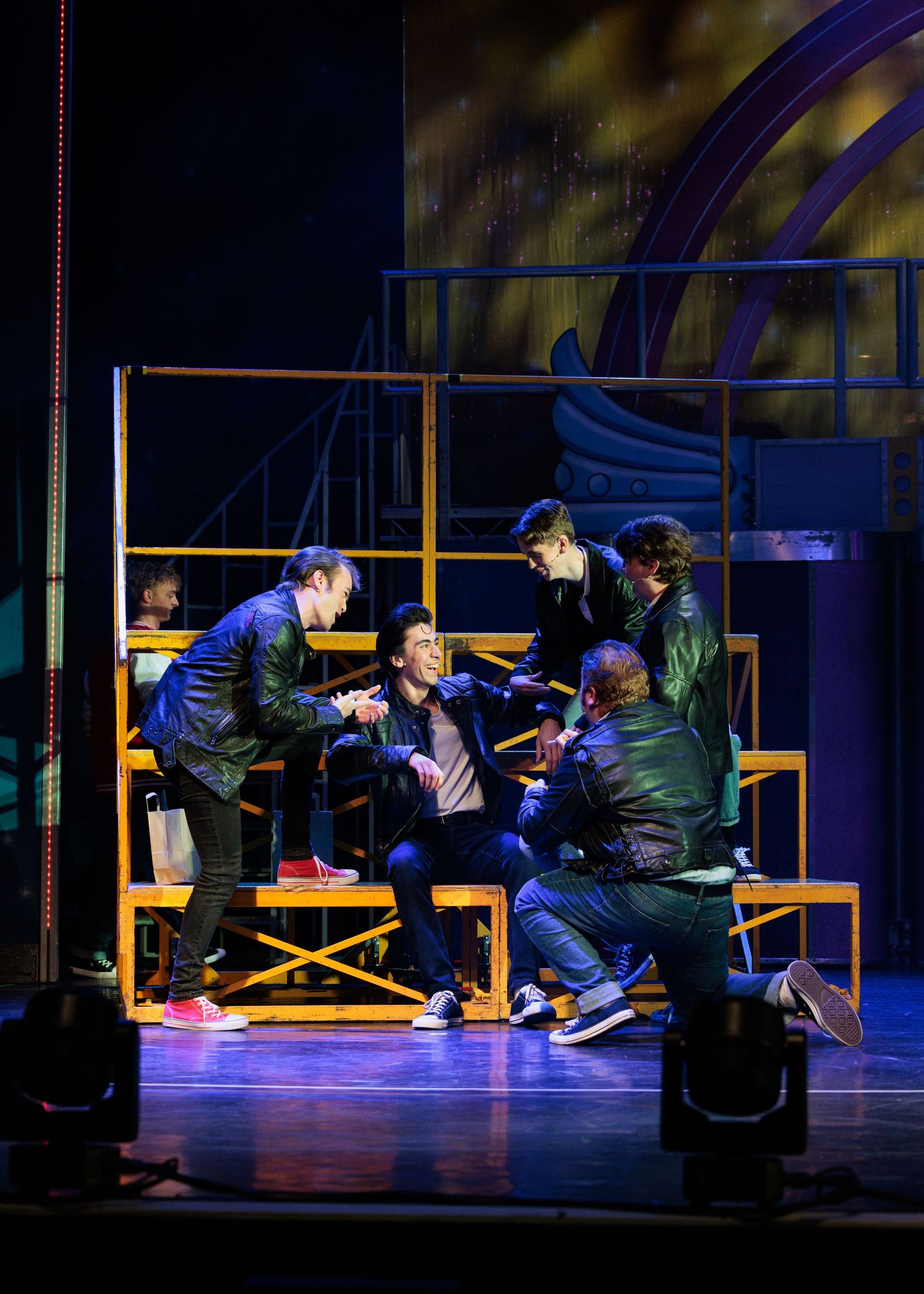 Dancers perform a dynamic sequence on yellow benches under blue stage lighting during a theatrical production.