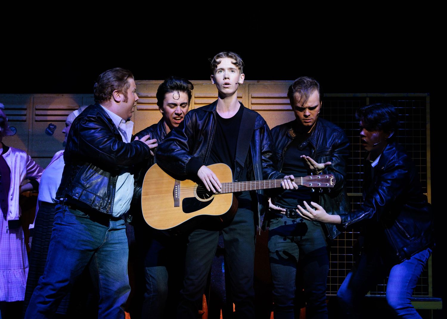 Musicians perform on stage with acoustic guitars under blue lighting during a theatrical performance.