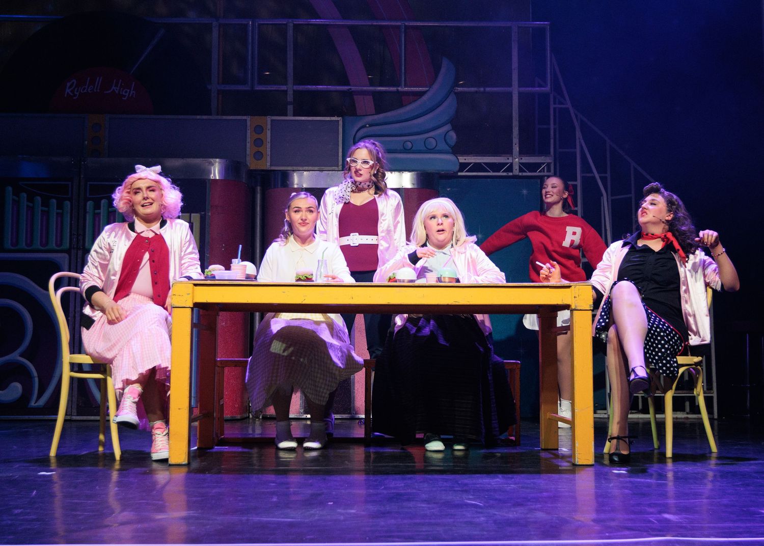 Stage performers sit on yellow benches during a theatrical scene with dramatic lighting.
