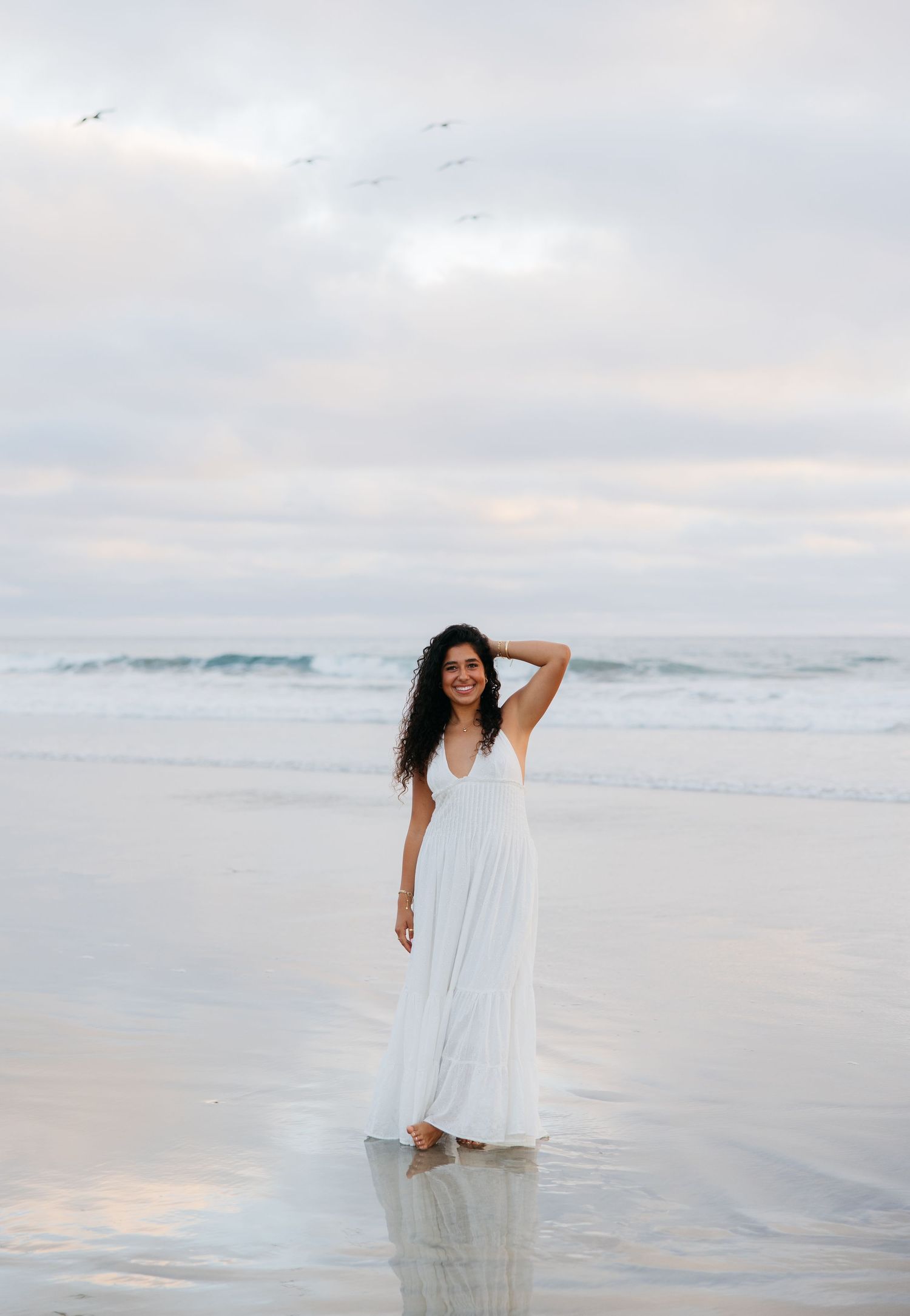 A figure in a flowing white dress poses gracefully in shallow ocean waters at sunset.