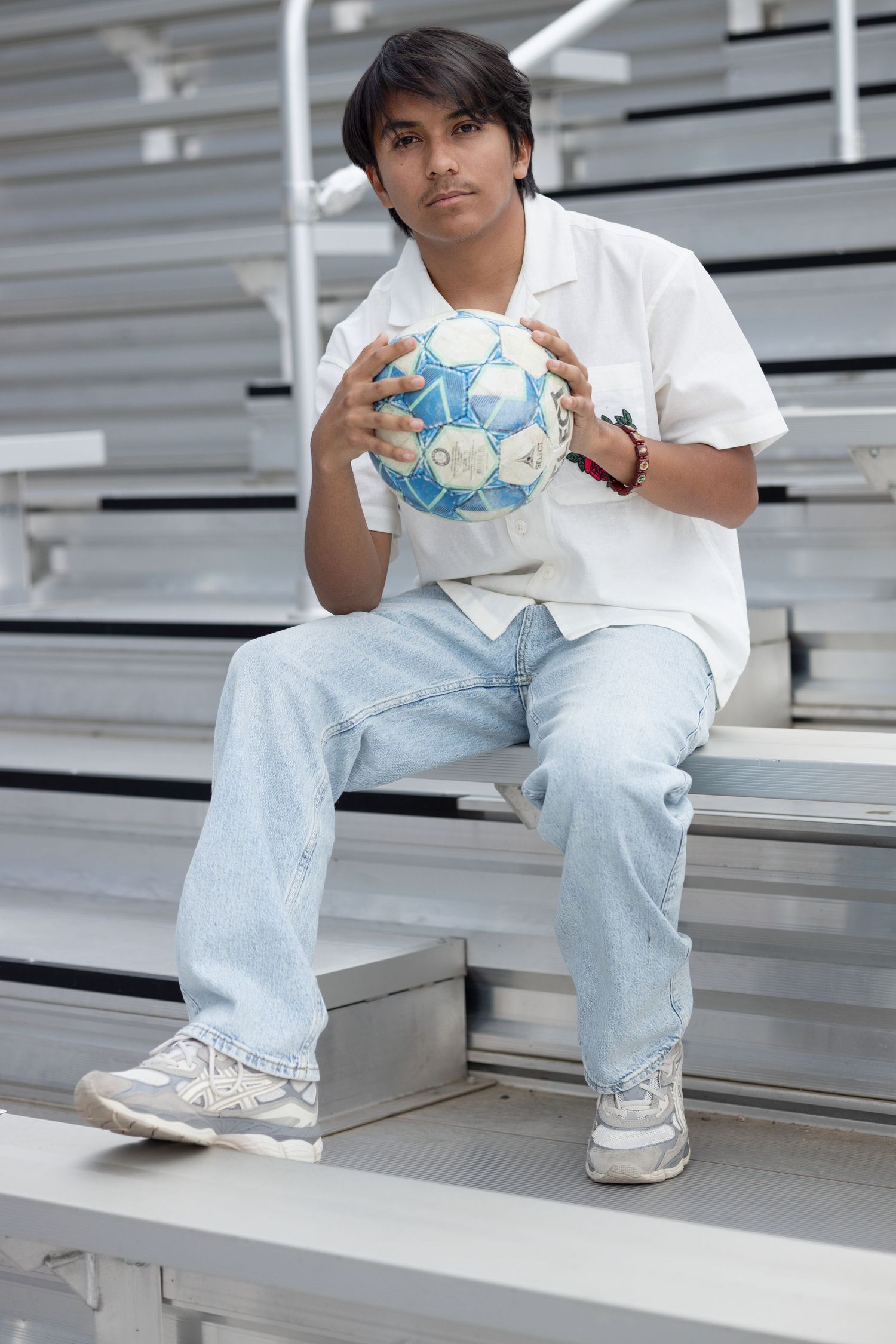 Person in white shirt and light jeans sitting on bleachers holding a blue and white soccer ball.