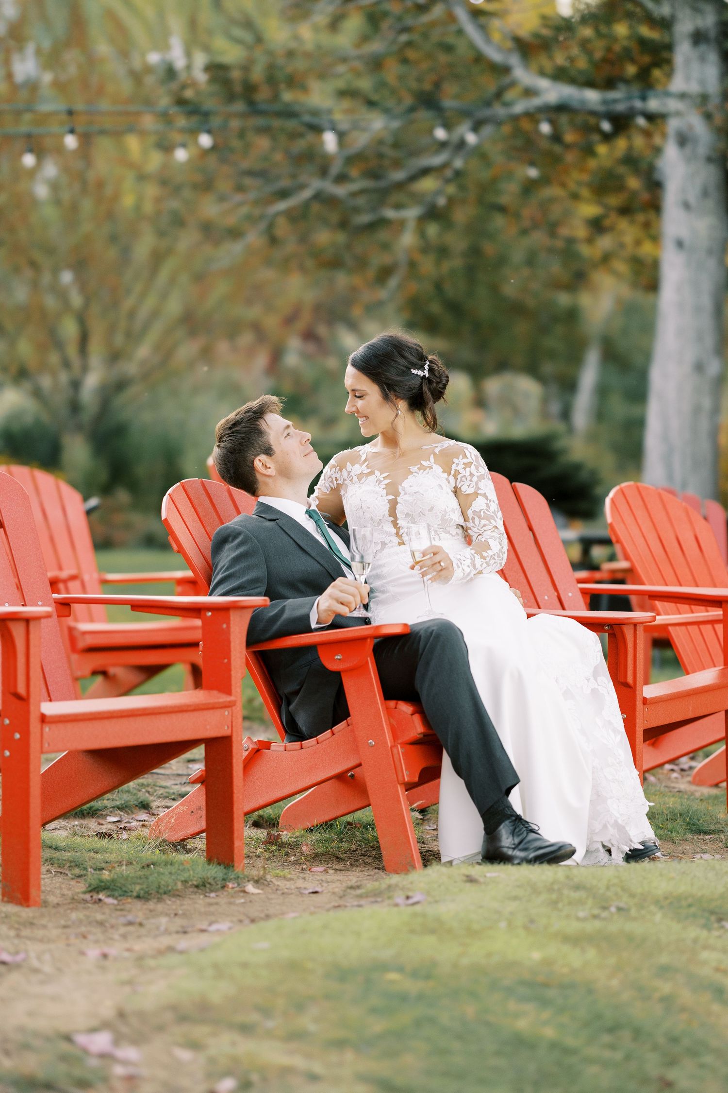 A romantic couple sits together in red Adirondack chairs during an outdoor wedding photoshoot.