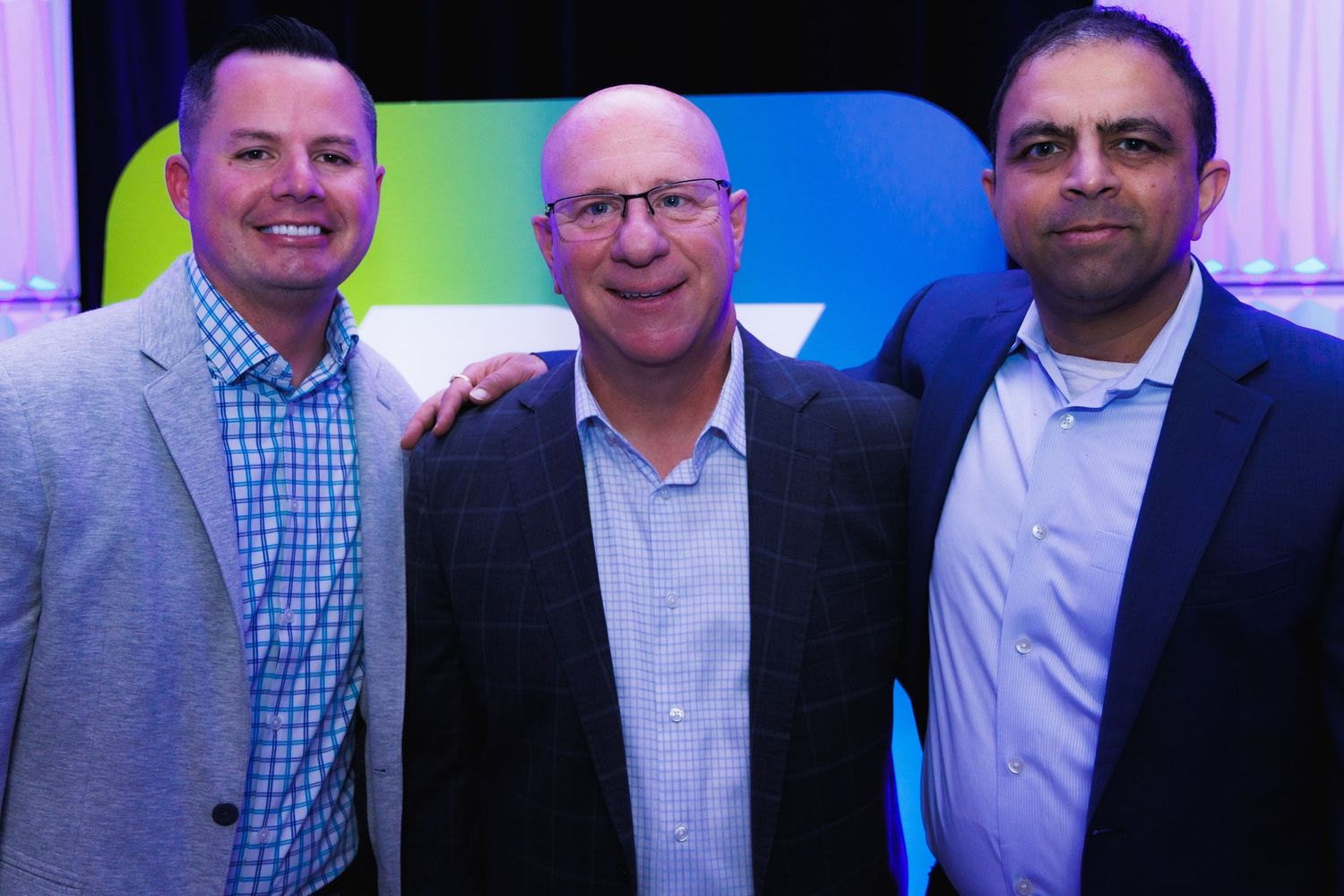 alt="Three corporate executives smiling together in front of Zips Car Wash branded backdrop at company conference"