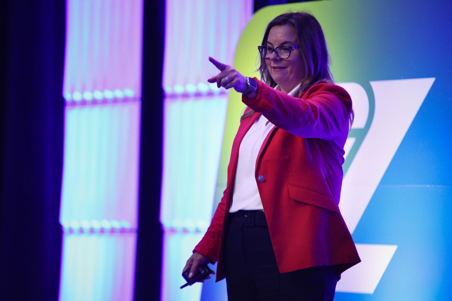alt="Female keynote speaker in red blazer presenting on stage with colorful purple and blue lighting at corporate event Orlando"
