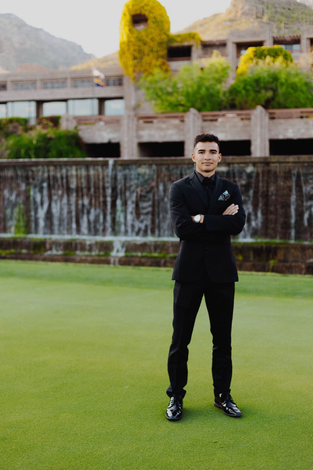 A person in black attire poses on a golf course with mountains in the background.