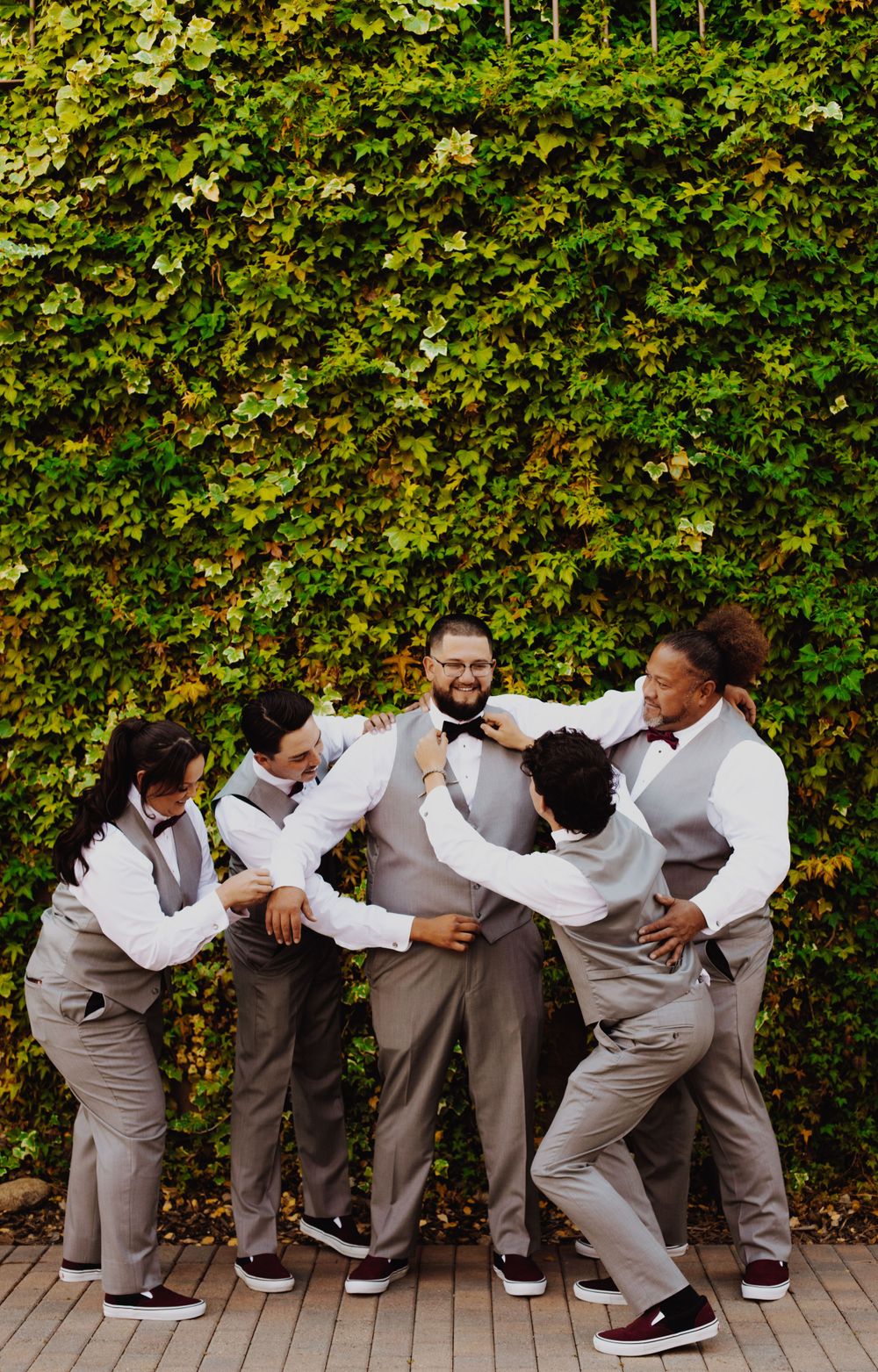 Groomsmen striking playful poses in a line against a green hedge backdrop.