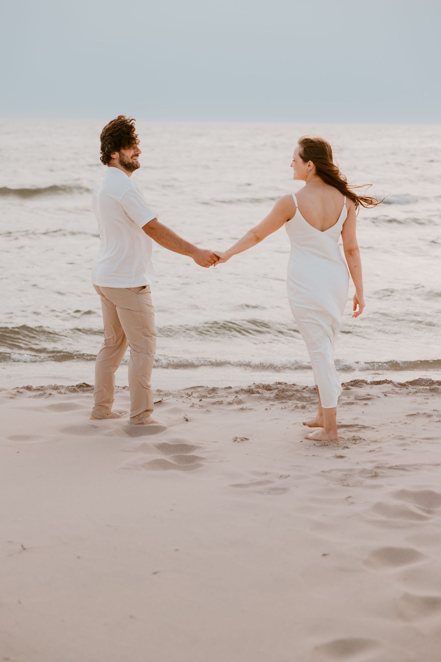 A couple in white outfits holding hands while walking along a beach at sunset with waves in the background. Michigan Wedding Photography