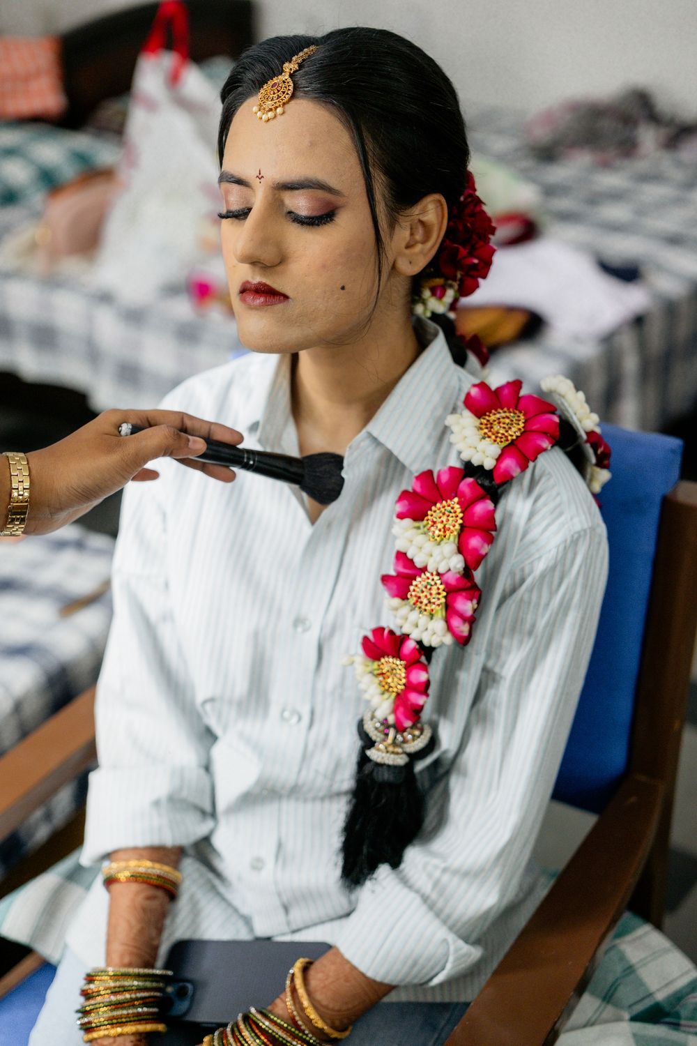 A person in traditional Indian bridal preparation wearing a white shirt with pink floral hair decorations and golden jewelry.