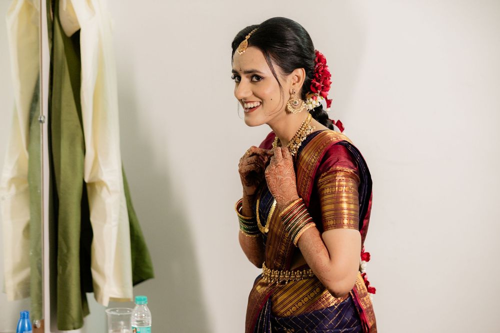 A traditionally dressed woman in burgundy silk sari with golden bangles and red floral hair accessories smiles gracefully indoors.