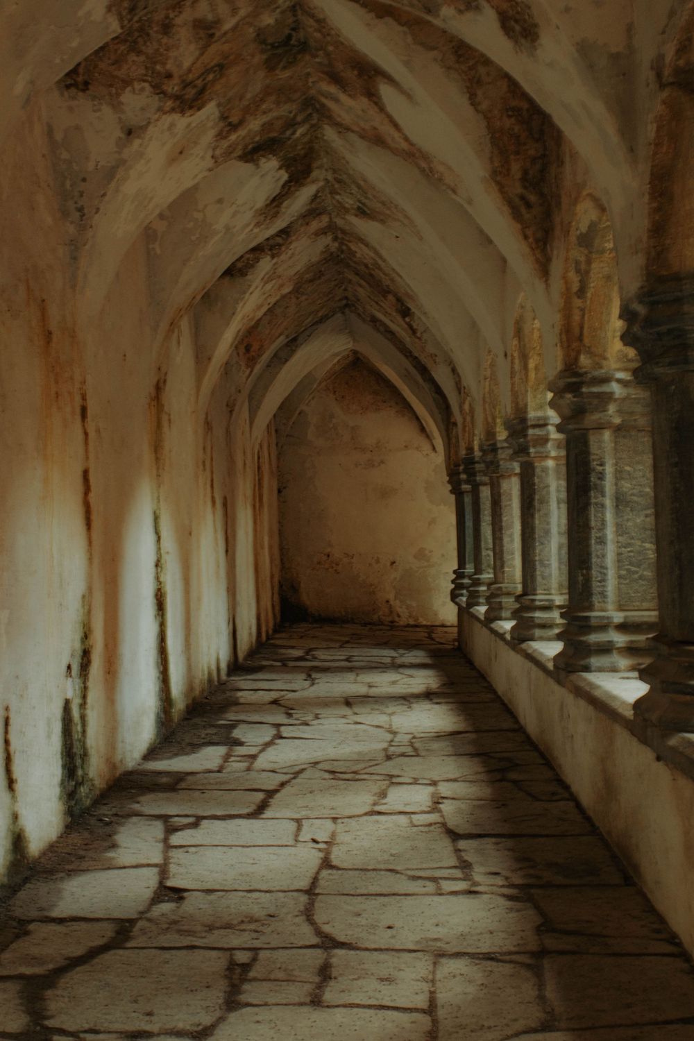 Ancient stone corridor with Gothic arched ceiling and weathered columns leads through a medieval monastery hallway.