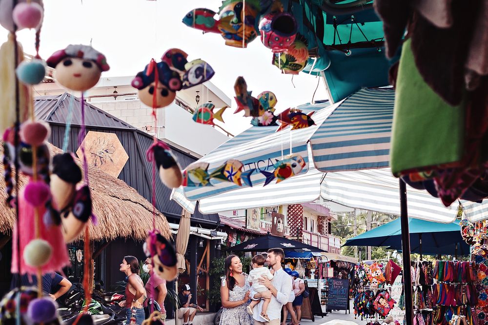 Colorful vendor umbrellas and stalls line a bustling outdoor market street with shoppers browsing merchandise.