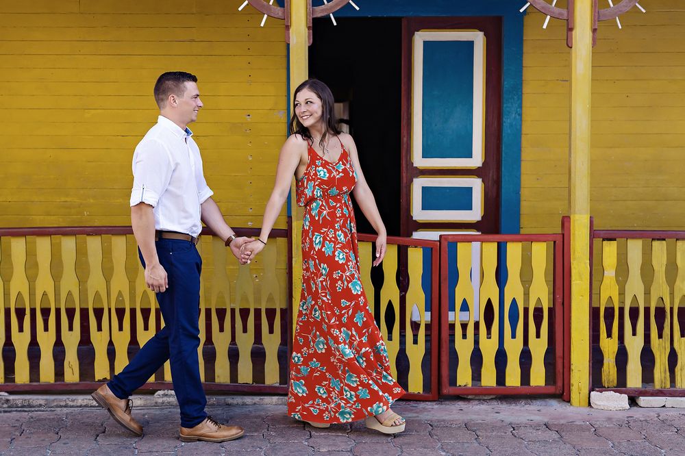 A couple holds hands while posing together on a porch with yellow walls and decorative porch trim.