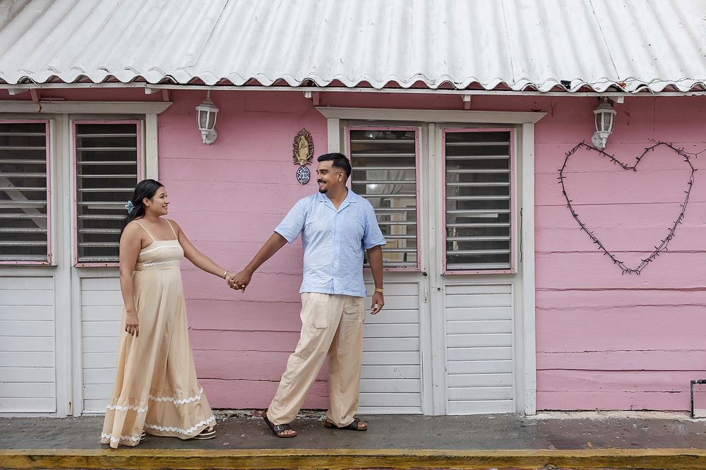A couple stands together holding hands against a pink wall with a heart design outside a building with shuttered windows.