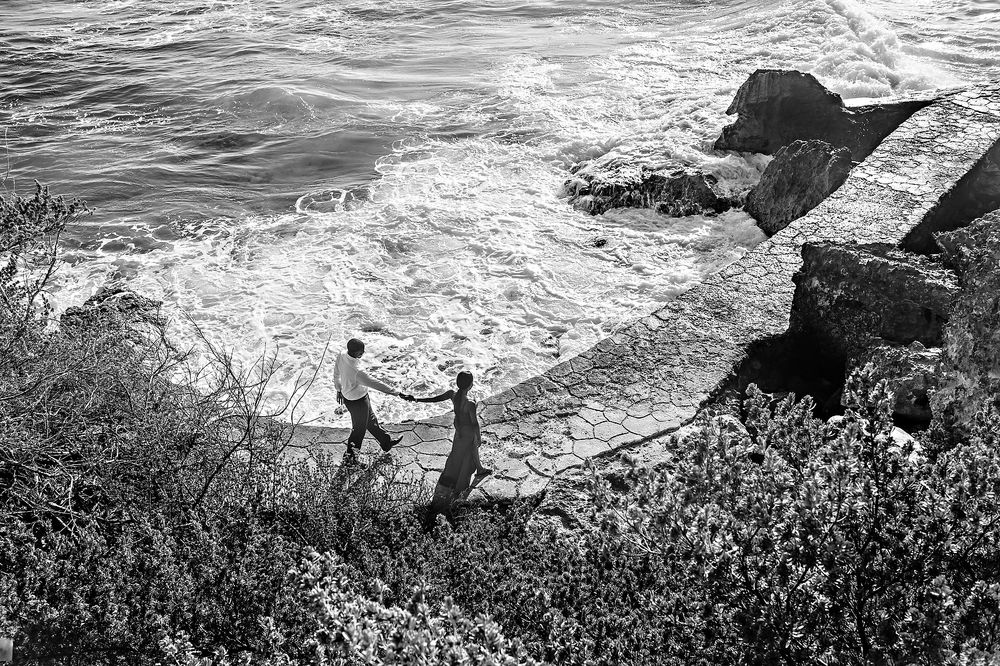 A black and white scenic coastal view shows two figures standing on a cliff edge overlooking crashing waves below.