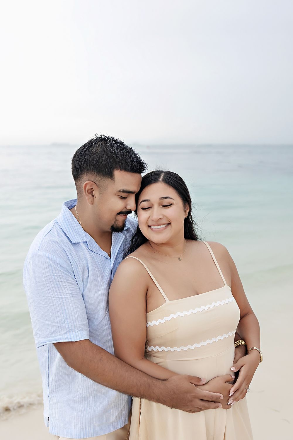 A romantic couple embraces on a peaceful beach with turquoise water and soft light in the background.