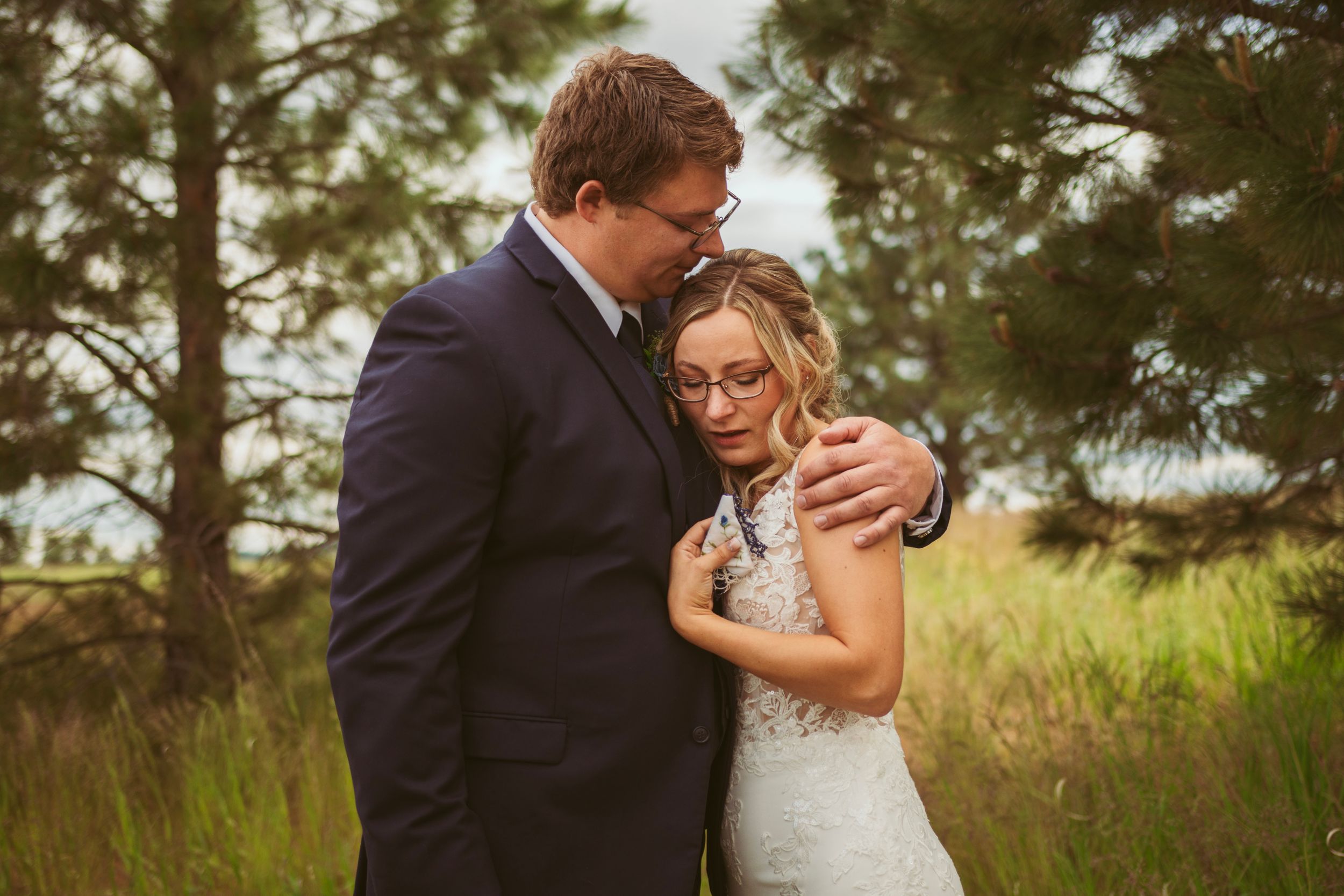 A tender embrace between a couple wearing formal attire standing in a meadow with pine trees in the background.