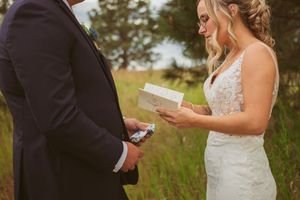 Couple holding and reading personal vows during outdoor summer wedding ceremony in tall grassy field.