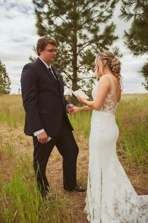 A couple in wedding attire shares a tender moment in a rustic outdoor setting with pine trees and tall grass.