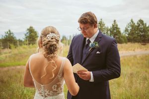 A couple exchanges vows during an outdoor wedding ceremony in a rustic field surrounded by evergreen trees.