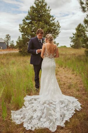 Elegant lace wedding gown with dramatic train photographed against natural grassland and evergreen backdrop.