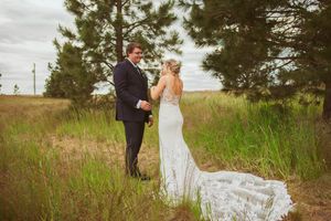 Lace wedding dress catches the sunlight as a couple interacts on a grassy hillside with pine trees.