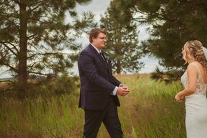 Wedding couple exchanges intimate gestures in a natural setting with evergreen trees and meadow grass.